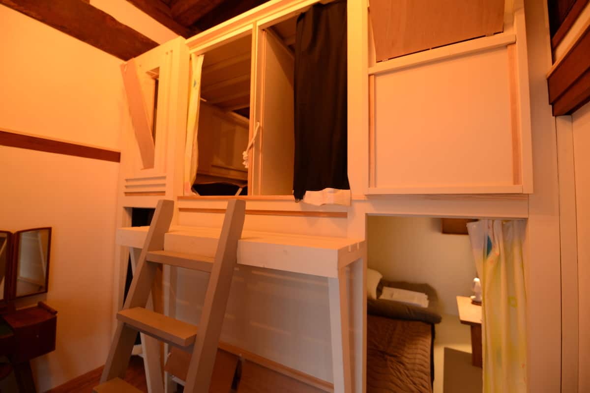 Een kamer met een stapelbed, een trapje, een klein nachtkastje en een raam met gordijnen.