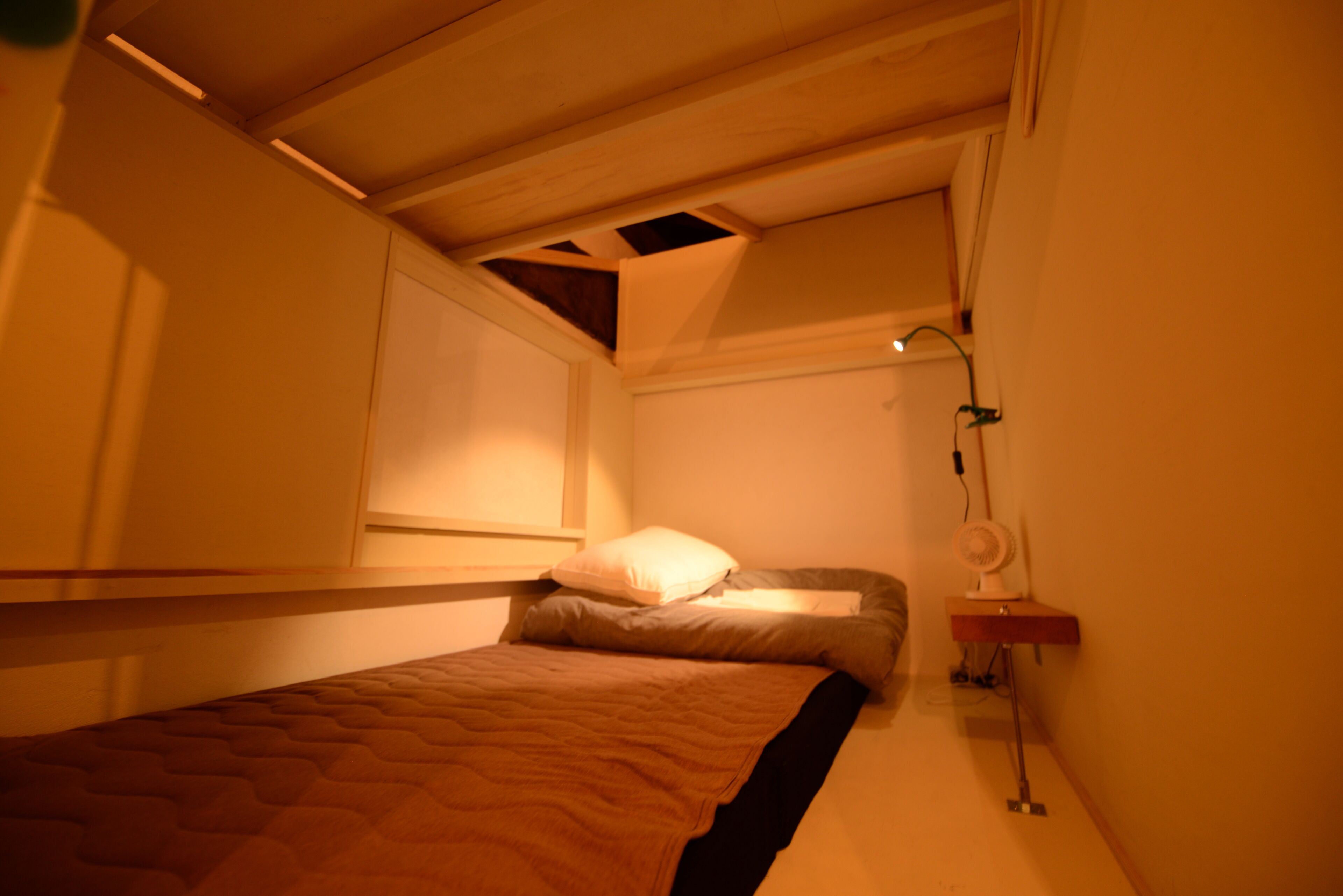 Een kleine kamer met een enkel bed, schuine wand, een raam en een nachtkastje met lamp.