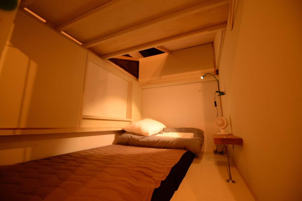 Een kleine kamer met een enkel bed, schuine wand, een raam en een nachtkastje met lamp.