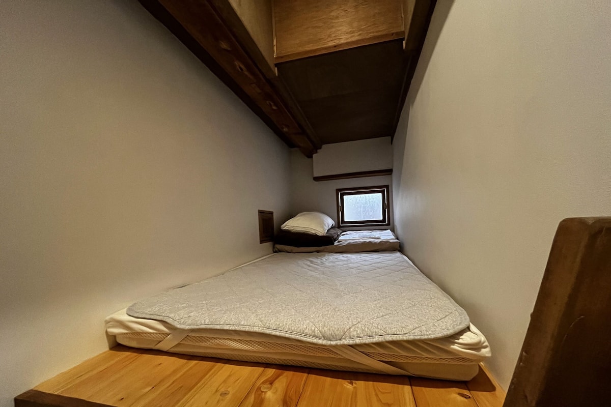 Een kleine, smalle kamer met een enkel bed, een houten tafel en een raam dat voor natuurlijk licht zorgt.