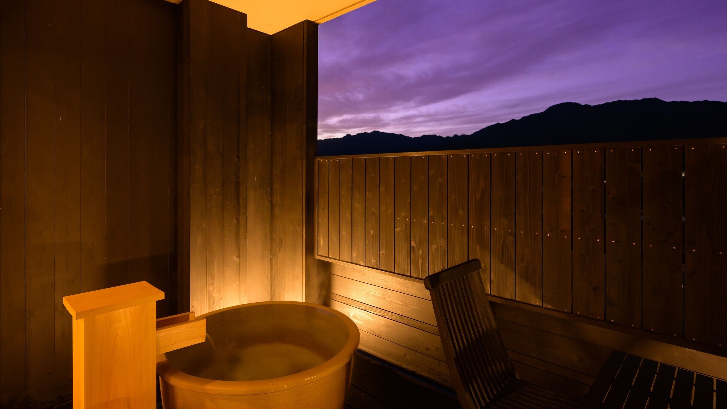 A wooden spa area with a hot tub and a chair, overlooking a mountain range at dusk.