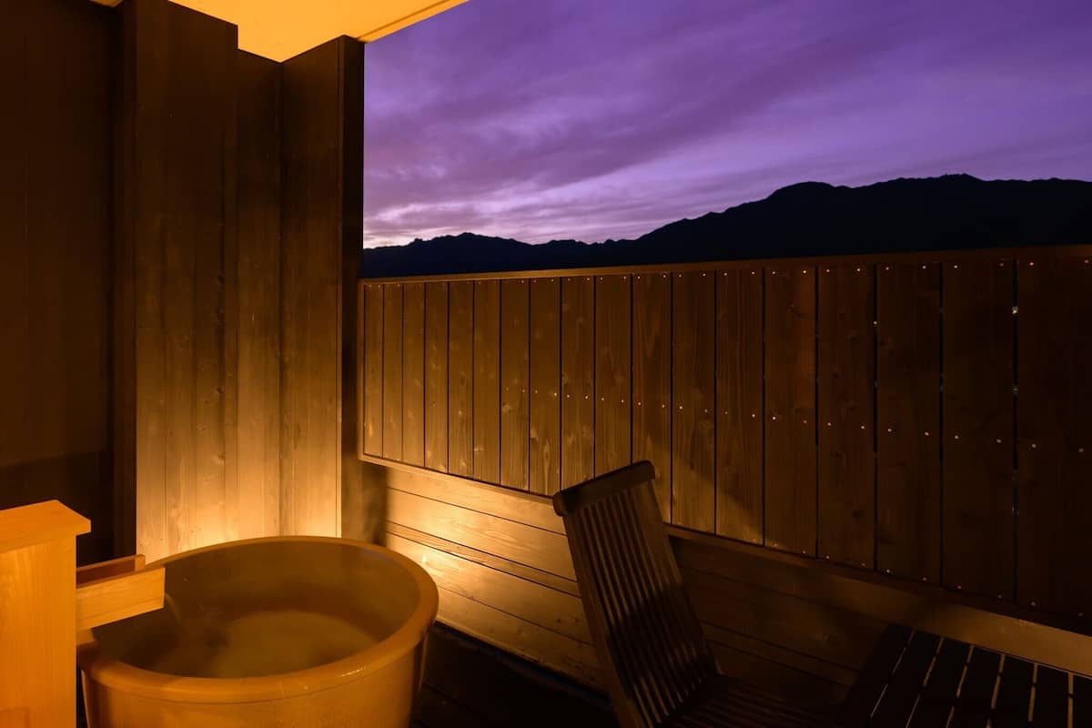 Un área de spa de madera con un jacuzzi y una silla, con vista a una cordillera al atardecer.