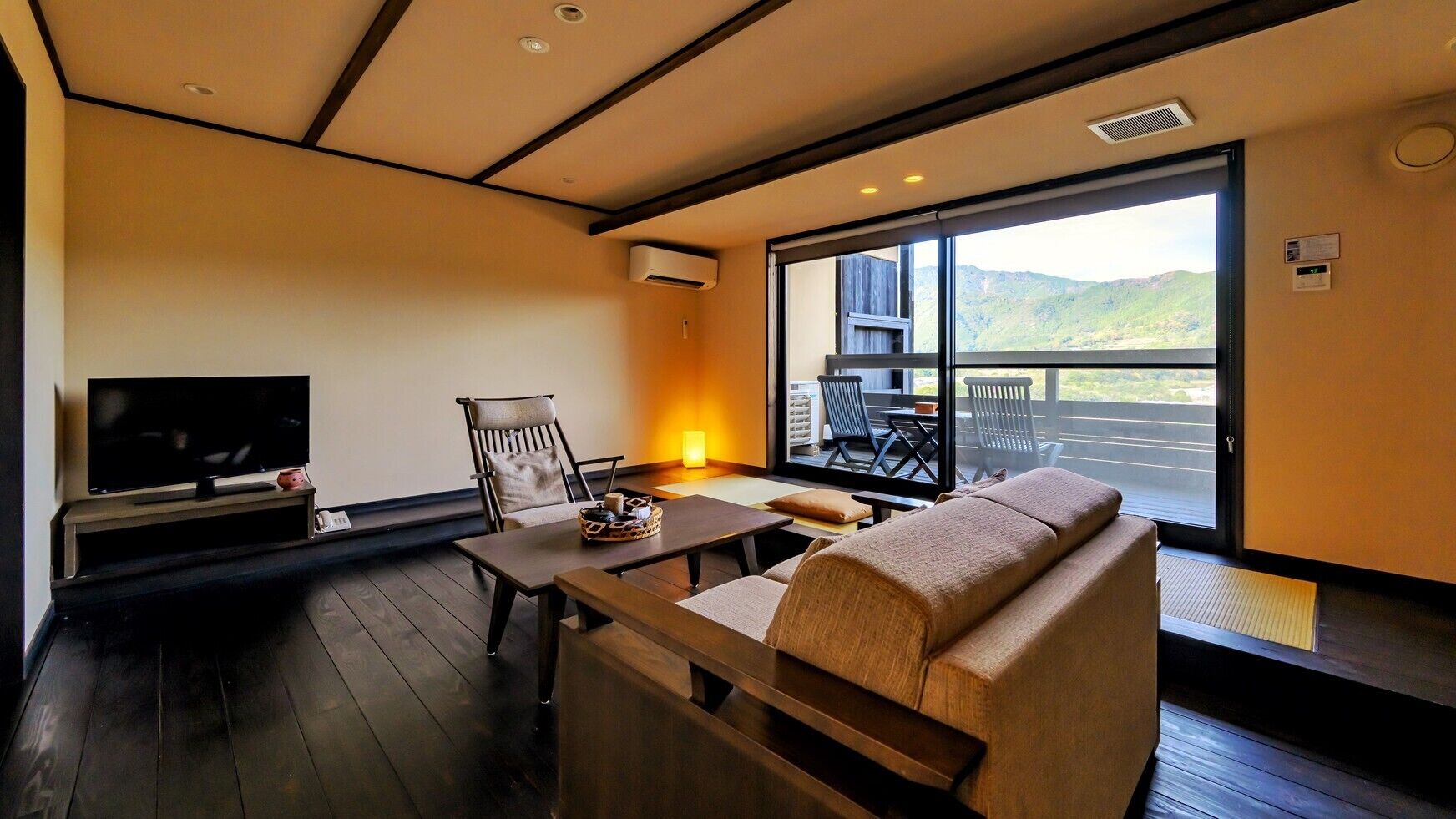 A modern living room with a sofa, a TV, a table with a decorative item, and a large window with a view of the outdoors.