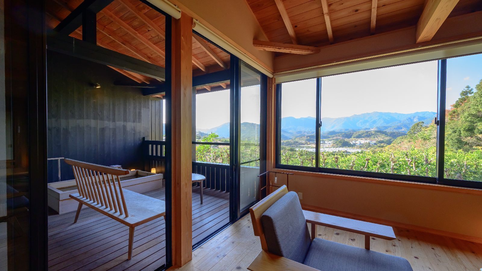 A room with a wooden deck, a sofa, a bench, and a large window offering a view of a vineyard and mountains.