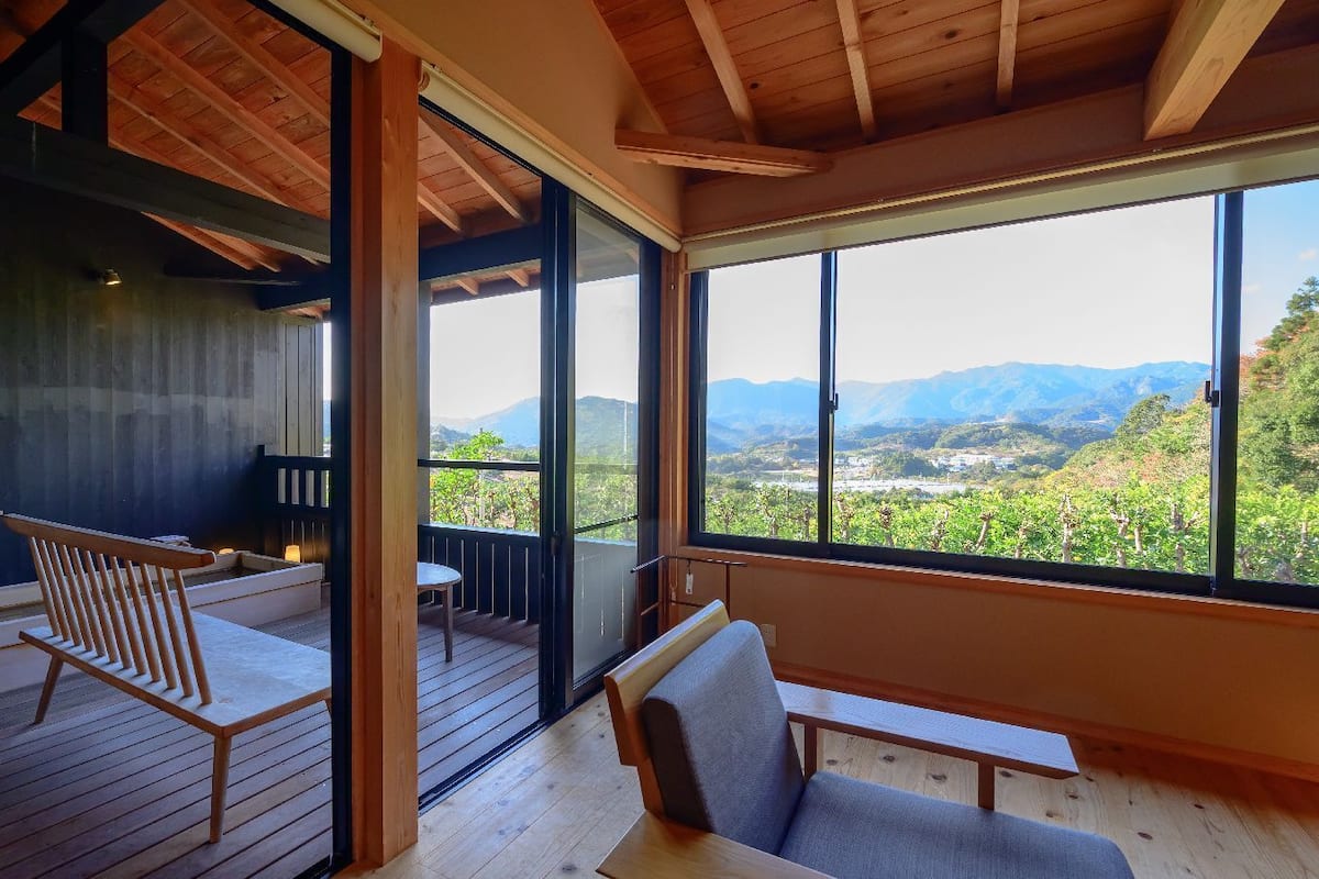 Una habitación con terraza de madera, sofá, banco y un amplio ventanal con vistas a un viñedo y montañas.