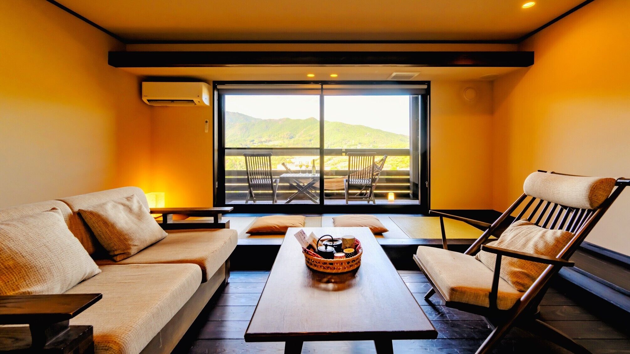 A room with a sofa, a chair, a table, and a view of a balcony with chairs and a table.