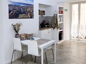 Un comedor con una mesa y sillas blancas, vistas a un paisaje urbano, y una zona de cocina con una cafetera y armarios.