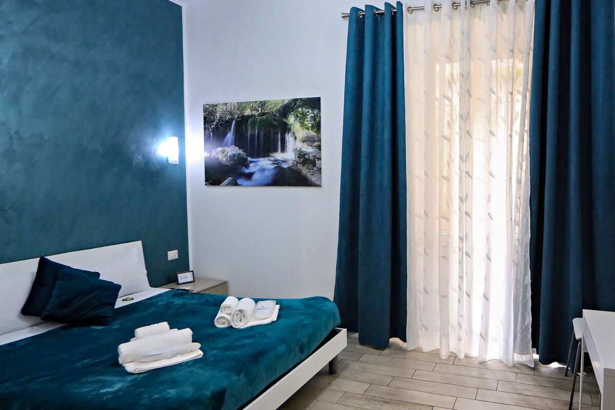 A bedroom with a bed, a nightstand, a wall-mounted picture, and a window with curtains.