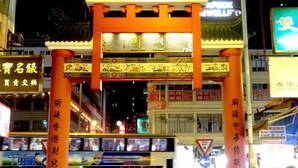 Un torii japonés tradicional por la noche, con letreros iluminados y comercios al fondo.