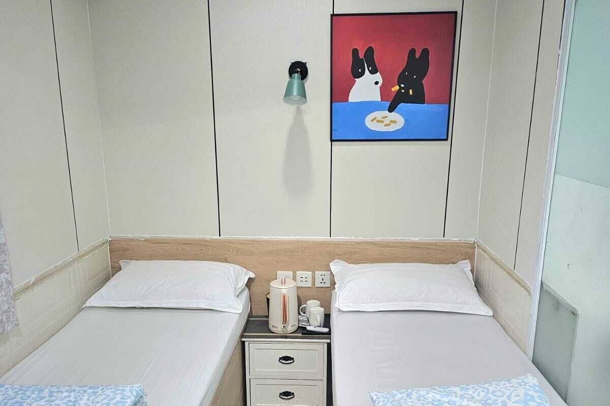 A twin room with two beds, a nightstand with a kettle, a wall clock, and a painting of dogs.