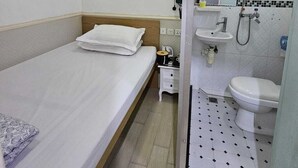 Habitación de hotel con una cama individual, una mesita de noche, un pequeño tocador, un espejo, un lavamanos y una ducha.