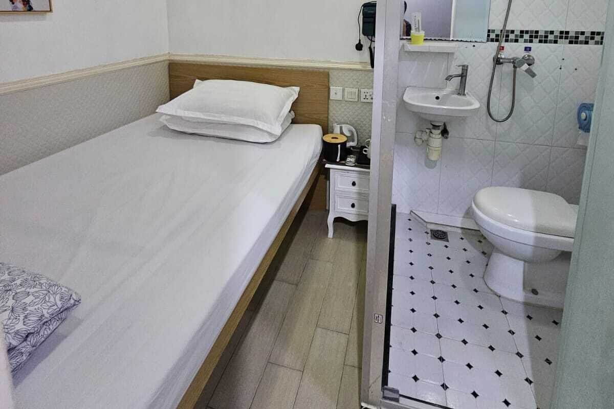 A hotel room with a single bed, a bedside table, a small nightstand, a mirror, a sink, and a shower.