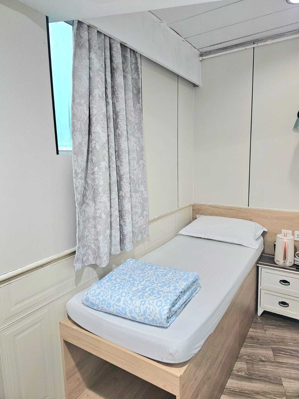 A single bed with a white mattress and a blue patterned blanket, a wooden headboard, a nightstand with a kettle, and a window with grey curtains.
