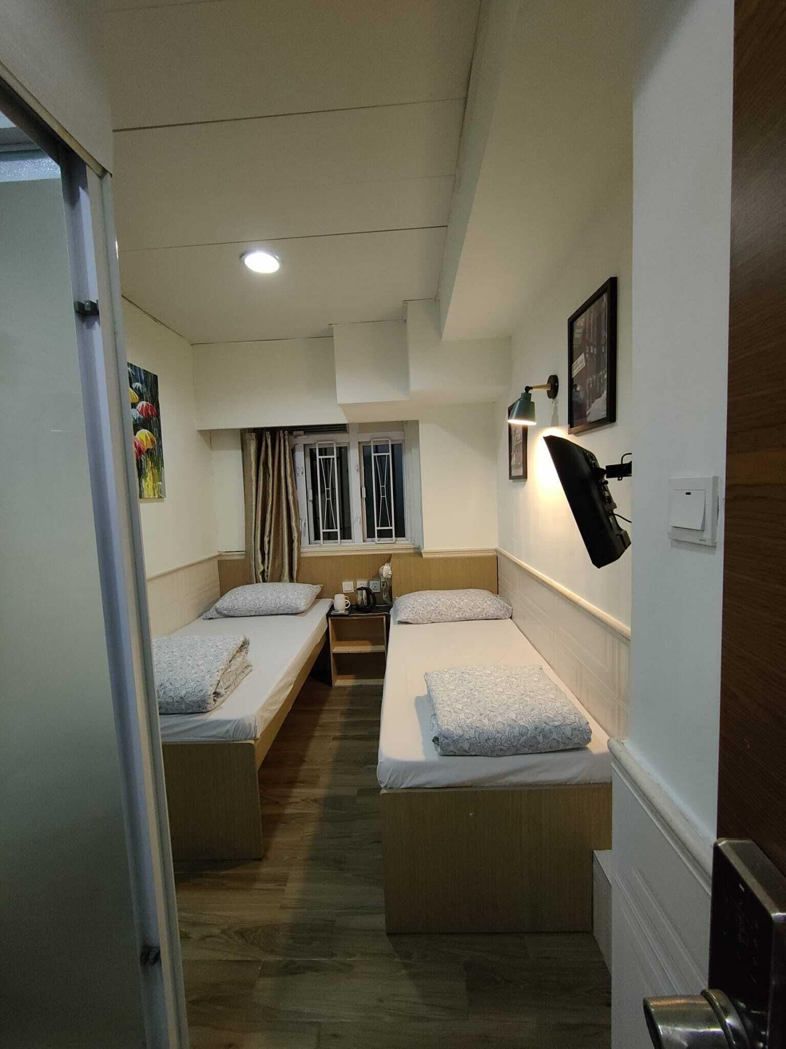 A compact hotel room with two single beds, a small table, a wall-mounted lamp, and a window with curtains.