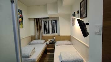 A compact hotel room with two single beds, a small table, a wall-mounted lamp, and a window with curtains.