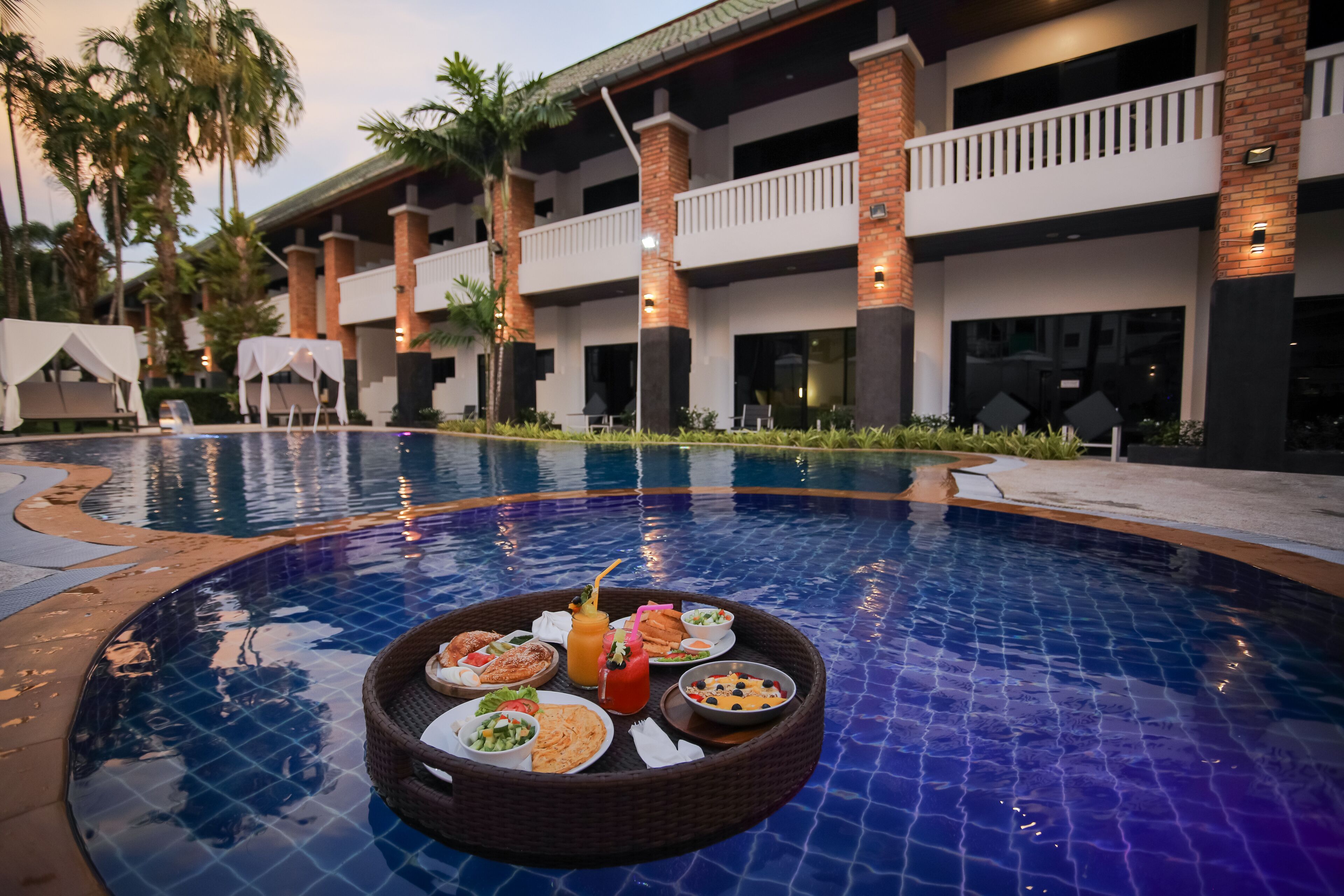 Mobiliario de comedor junto a la piscina, con diversos platos y bebidas, rodeado por un edificio con múltiples balcones y palmeras.