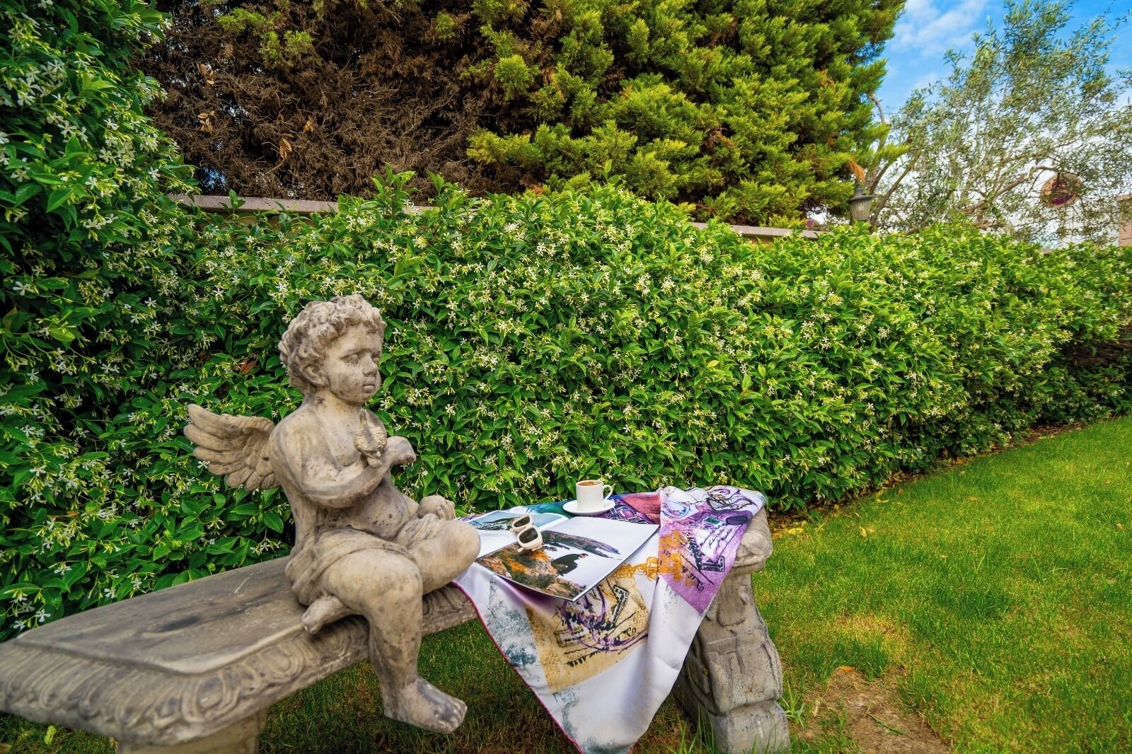 A statue of a cherub sitting on a bench with a table covered by a colorful cloth, surrounded by lush greenery.