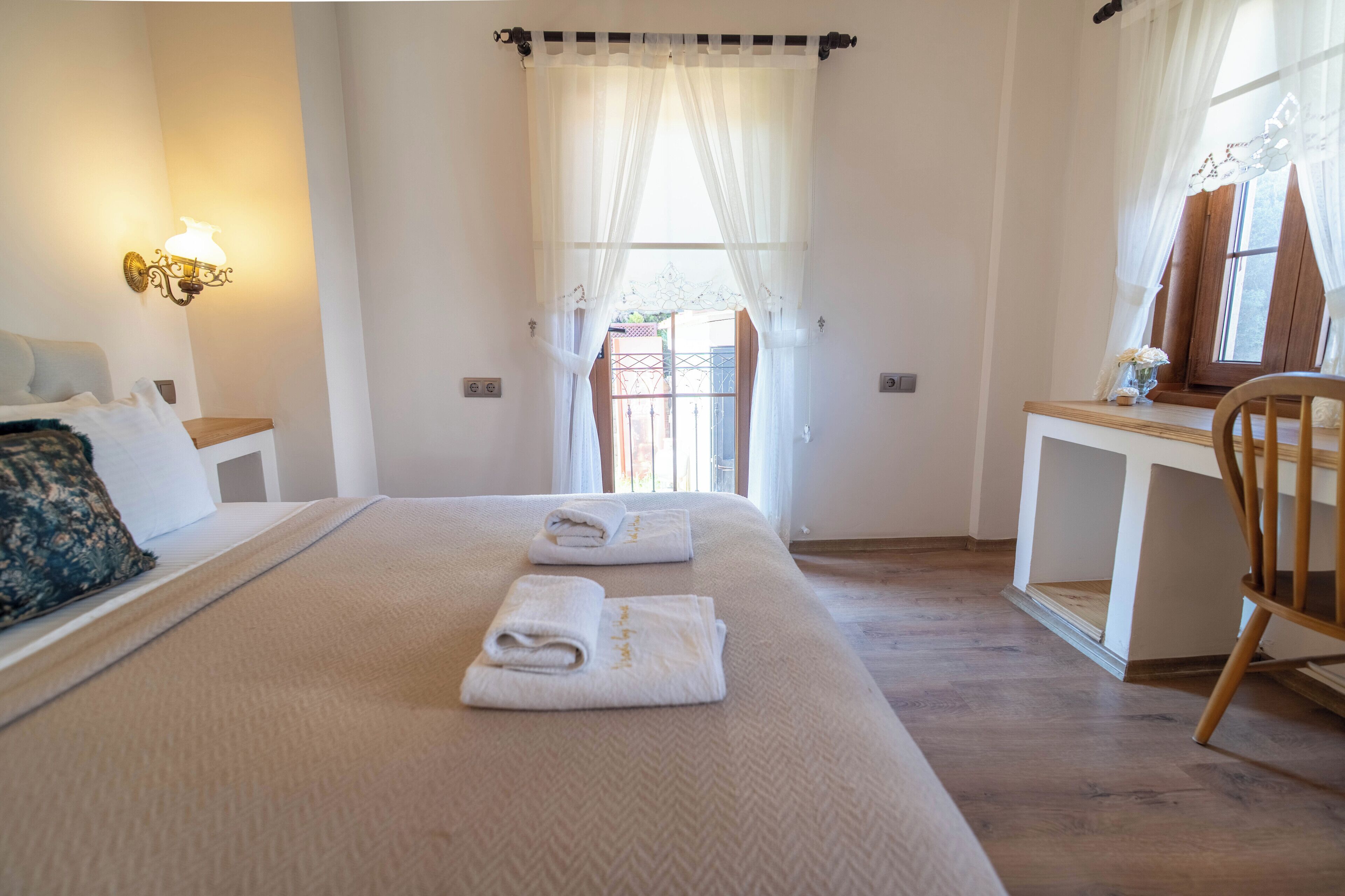 A hotel room with a bed, bedside table, lamp, window with curtains, and a desk with a chair.