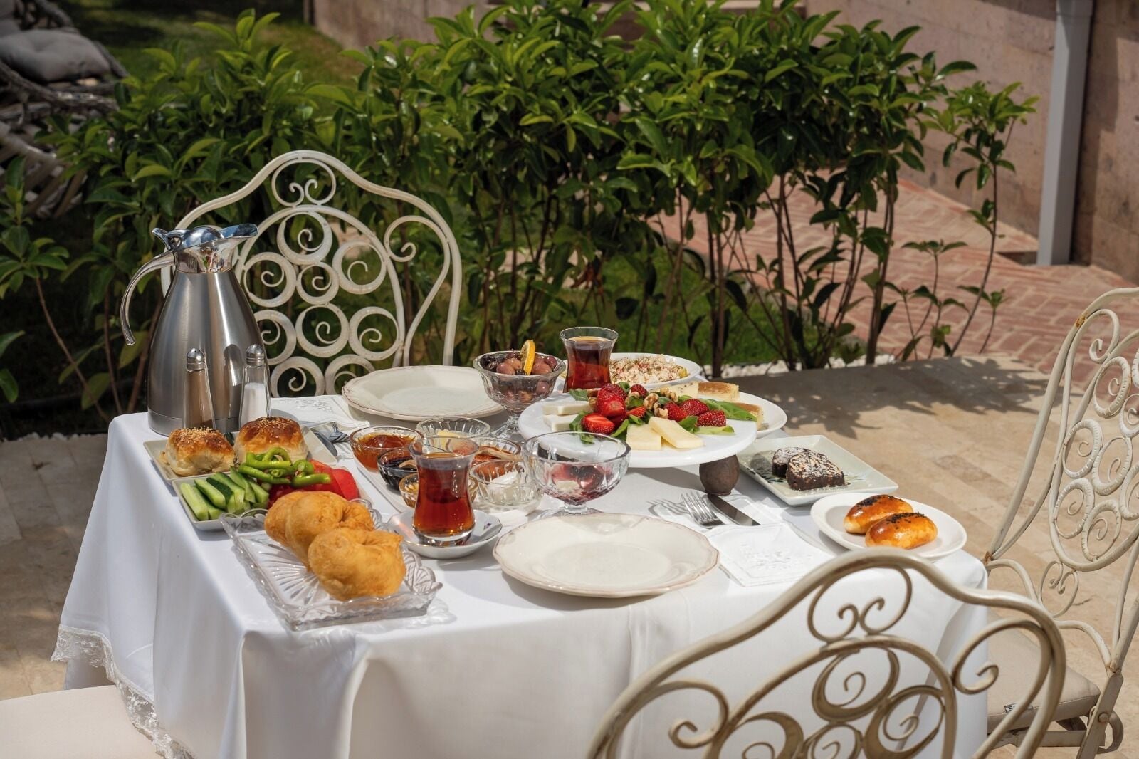A table set outdoors with a variety of dishes, including bread, vegetables, and desserts, and a teapot.