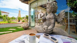 A stone statue of a seated figure with a cup and sunglasses on a table, set against a backdrop of a glass building and outdoor seating area. - Cunda Vrodi By House (Ayvalik)