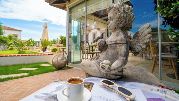 A stone statue of a seated figure with a cup and sunglasses on a table, set against a backdrop of a glass building and outdoor seating area. - Cunda Vrodi By House (Ayvalik)
