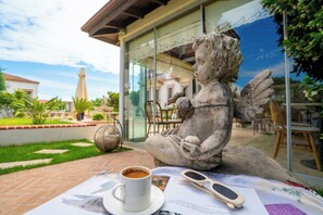 Una estatua de piedra de una figura sentada con una taza y gafas de sol sobre una mesa, frente a un edificio de cristal y una zona de estar al aire libre.