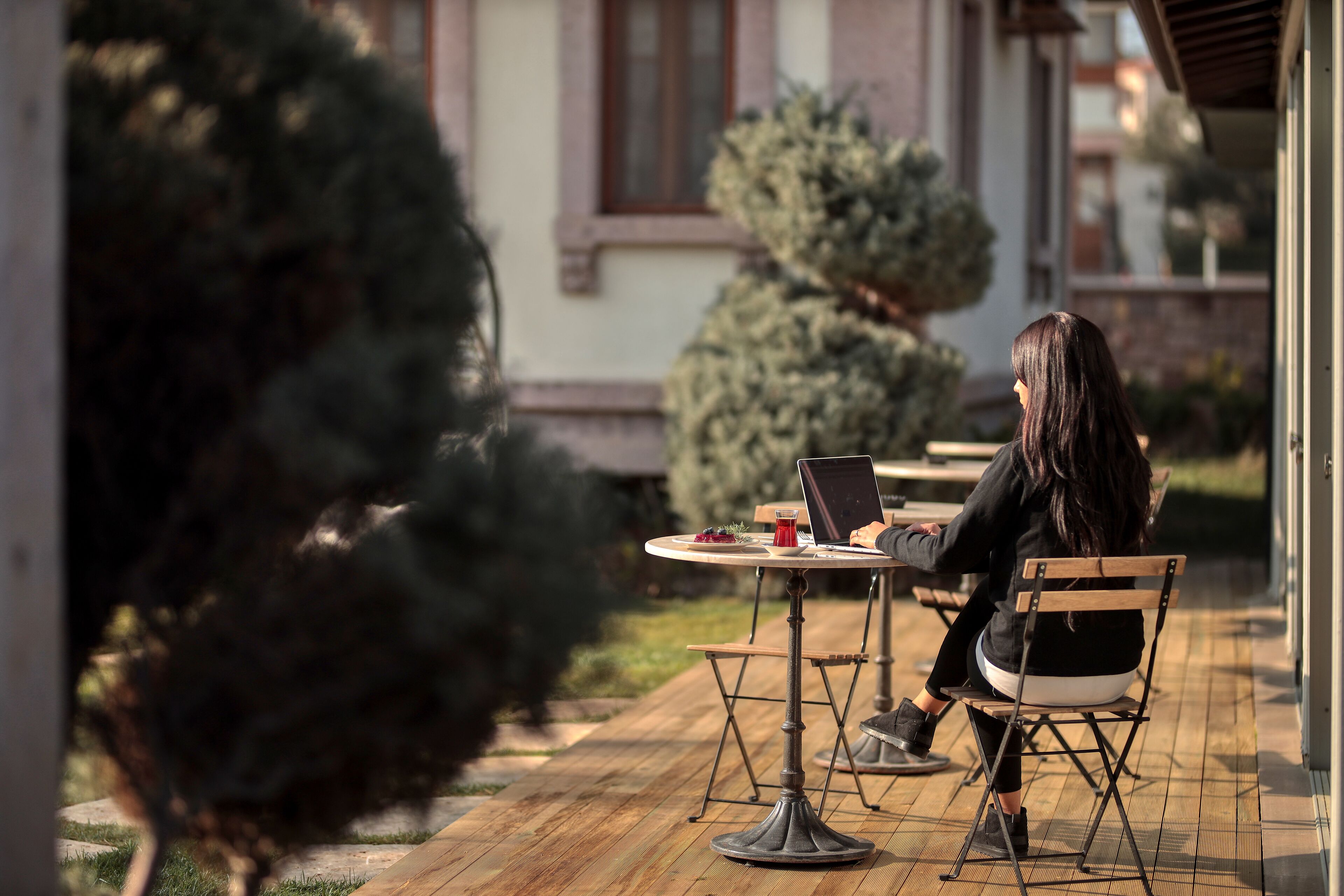 A person seated at an outdoor table with a laptop, surrounded by greenery and a building in the background.