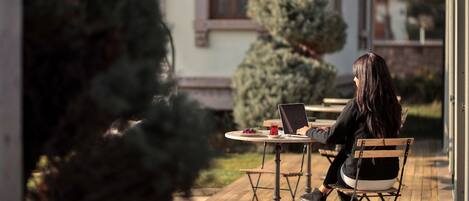 A person seated at an outdoor table with a laptop, surrounded by greenery and a building in the background.