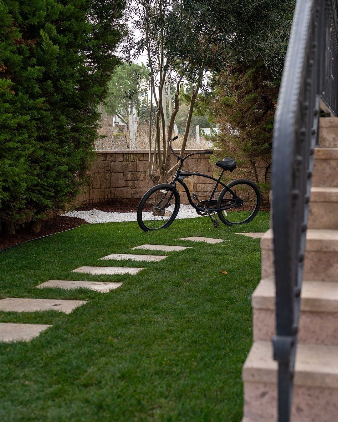 Una bicicleta aparcada en un sendero de piedra en un jardín, con un muro de piedra y árboles al fondo.
