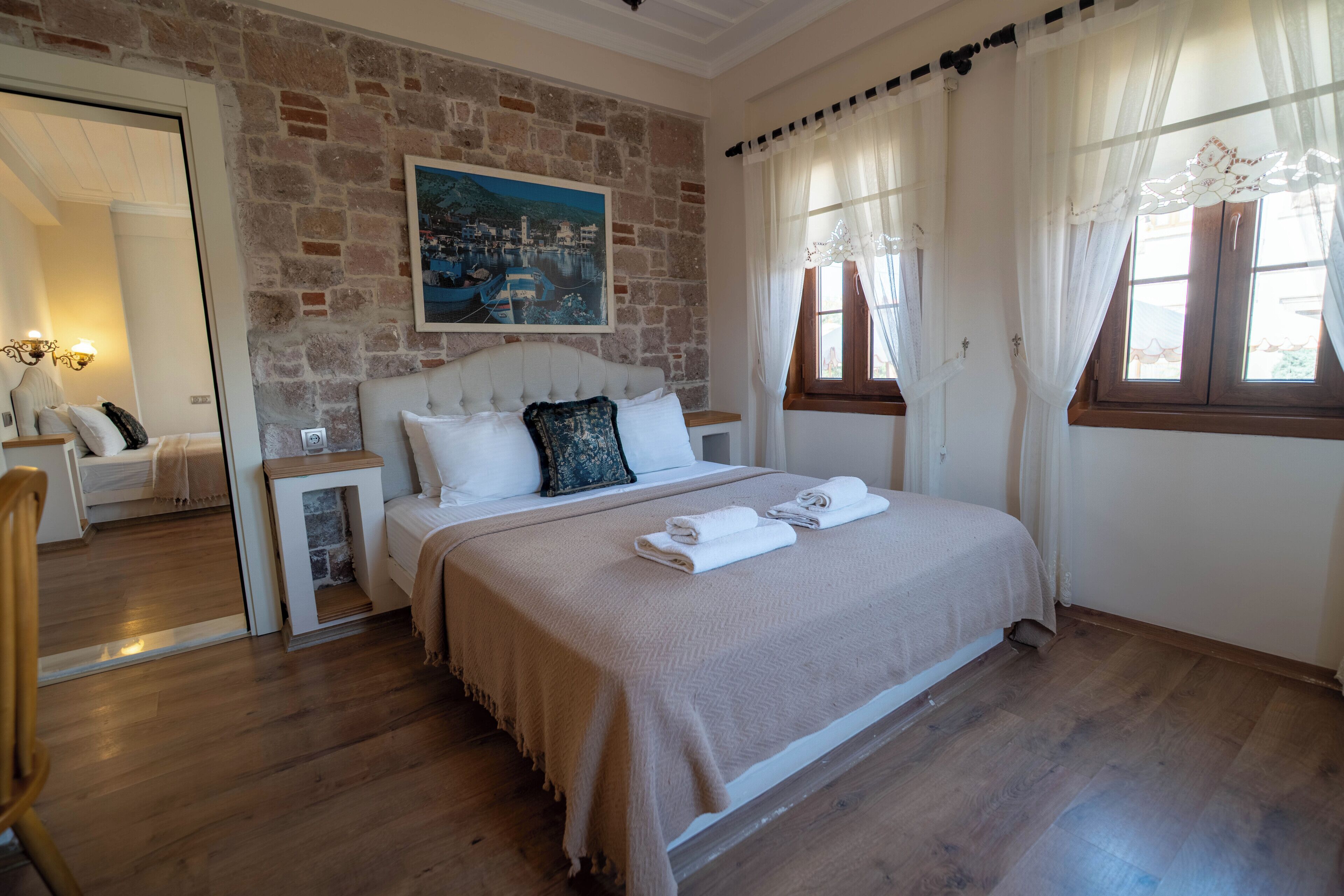 Un dormitorio con pared de piedra, una cama con cabecera, una mesita de noche y un espejo, además de una ventana con cortinas.