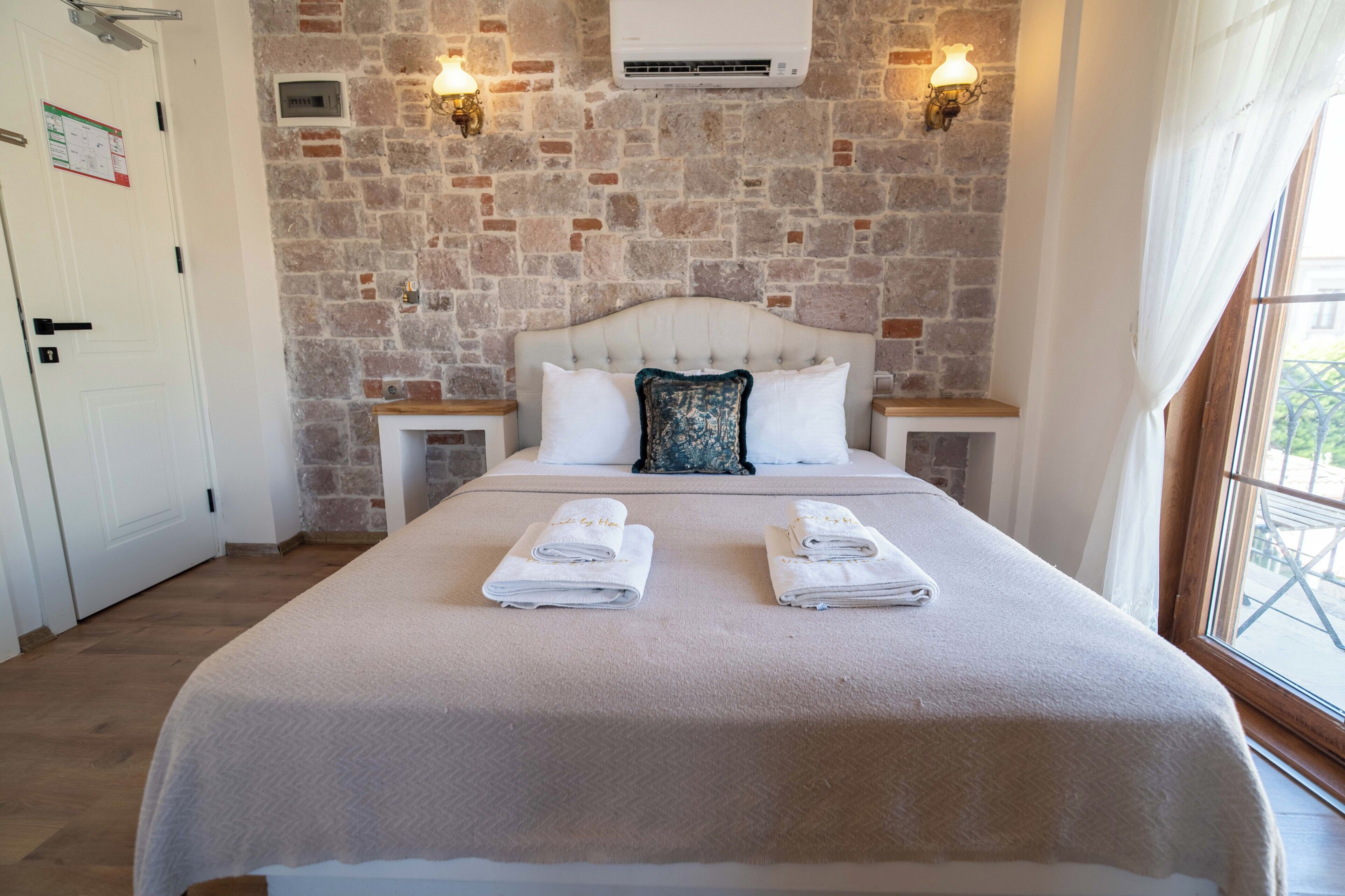 Une chambre d’hôtel avec un mur en pierre, un lit doté d’une tête de lit blanche, deux tables de chevet et une fenêtre avec des rideaux.
