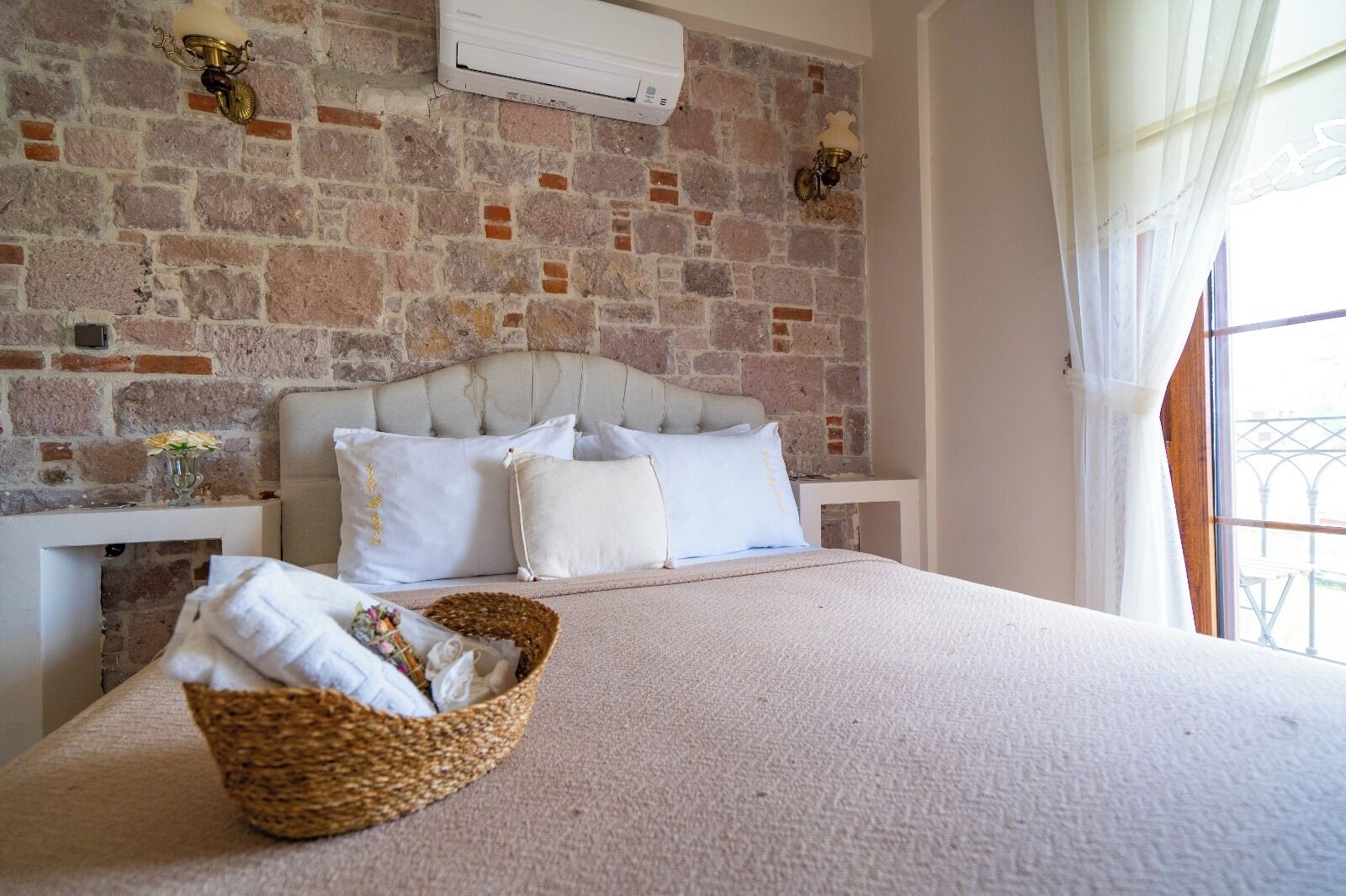 Une chambre avec un mur en briques, un lit avec une literie blanche, un panier en osier et une fenêtre avec des rideaux.