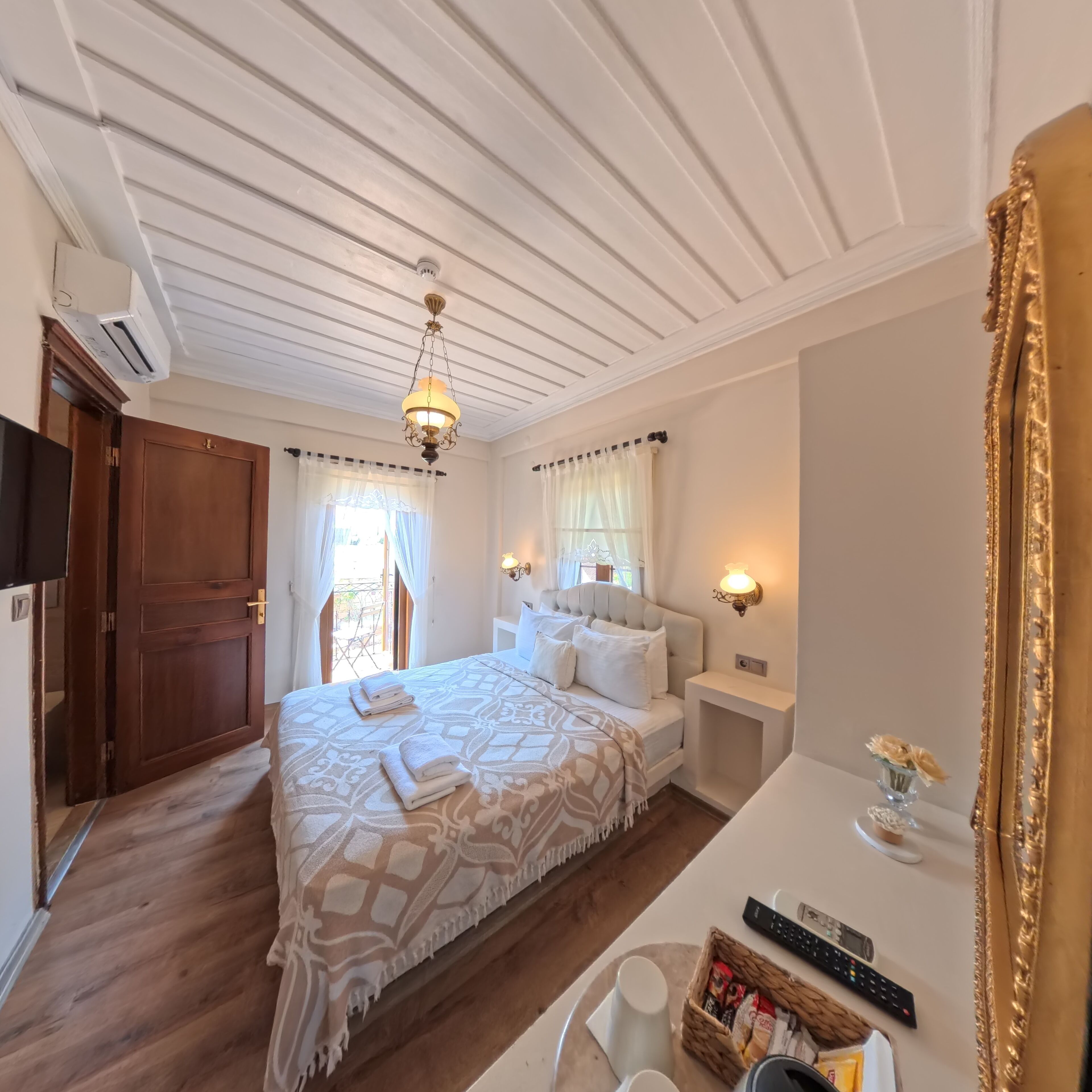 A hotel room with a large bed, a bedside table with a lamp, a small table with a vase and remote controls, and a large mirror.