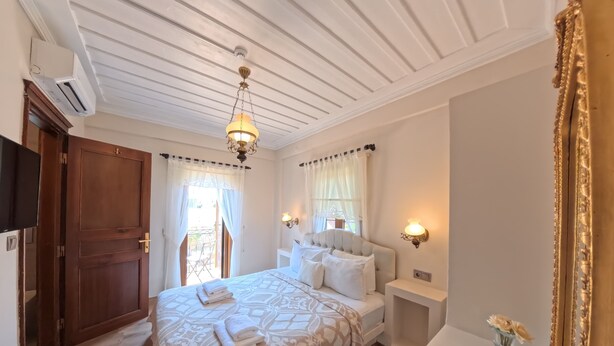 A hotel room with a large bed, a bedside table with a lamp, a small table with a vase and remote controls, and a large mirror. - Cunda Vrodi By House (Ayvalik)