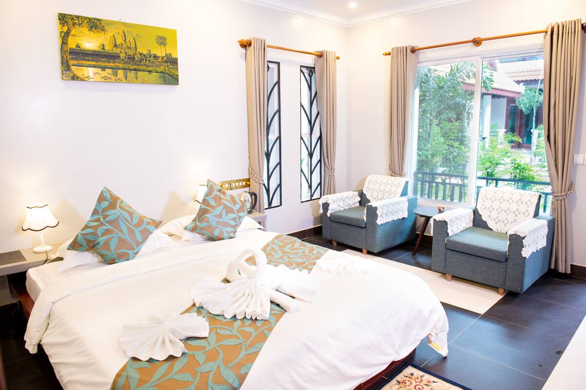 A hotel room with a bed, two armchairs, a small table, and a view of greenery outside.