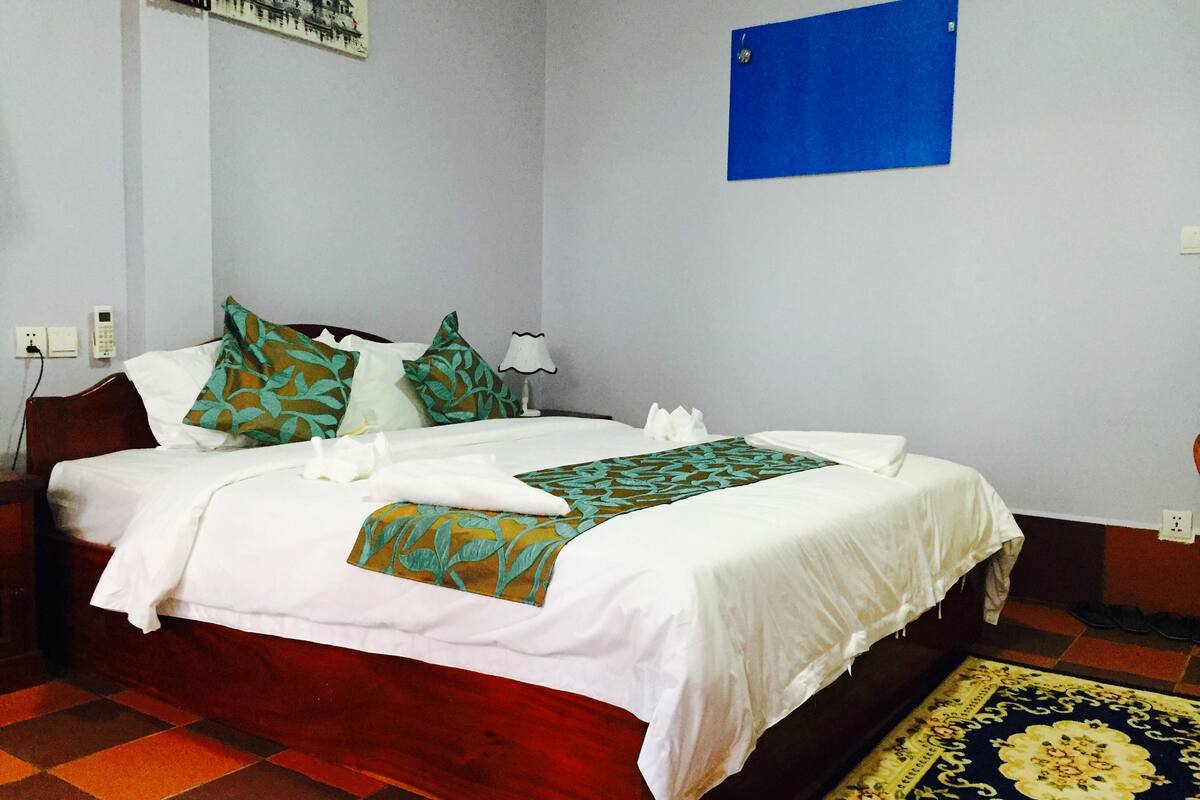 A neatly made bed with white linens and decorative pillows, a blue wall hanging, and a patterned rug.