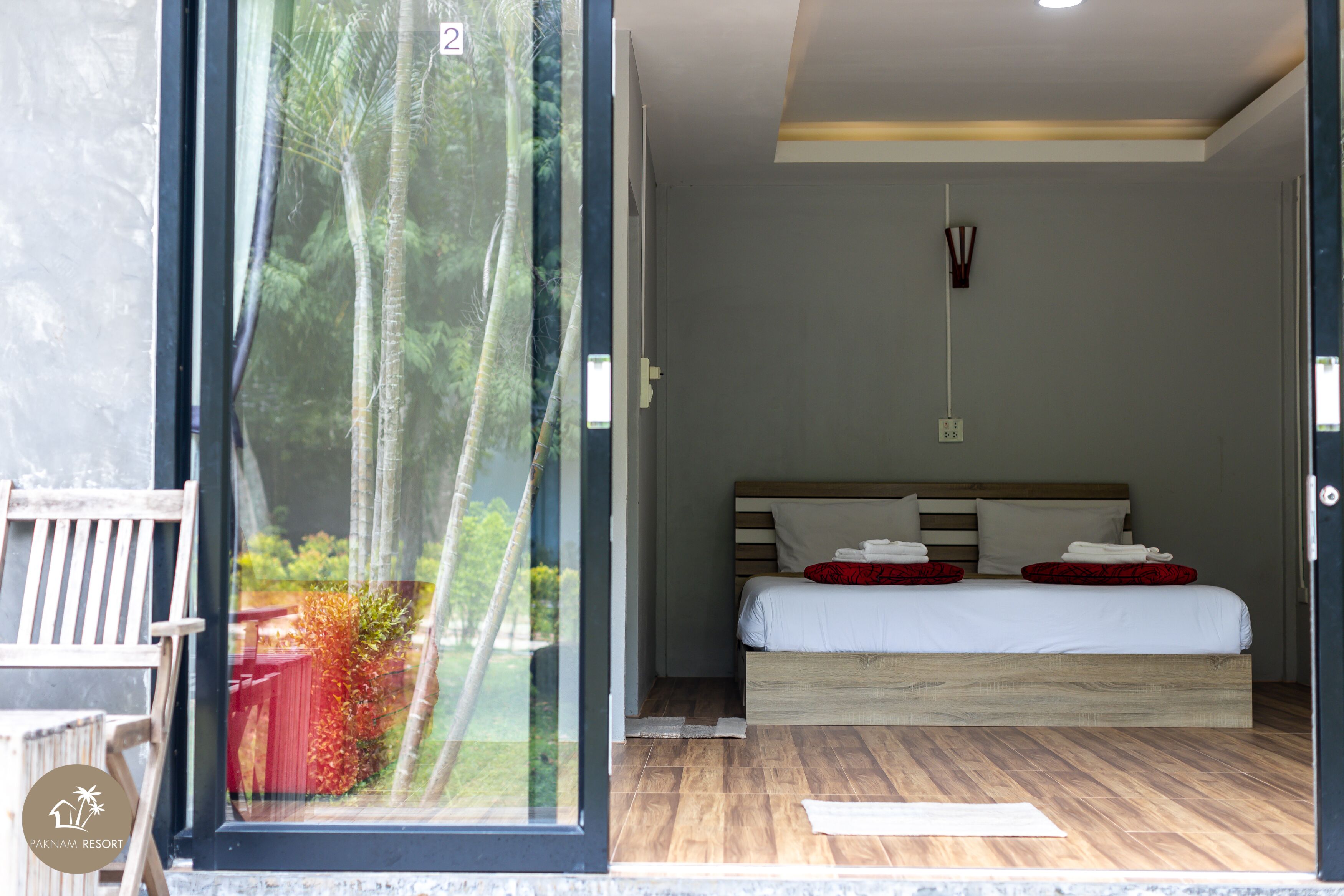 Quarto com uma grande janela, cama com roupa de cama branca e almofadas vermelhas, cabeceira de madeira e uma pequena mesa de cabeceira.