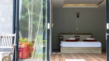Quarto com uma grande janela, cama com roupa de cama branca e almofadas vermelhas, cabeceira de madeira e uma pequena mesa de cabeceira.