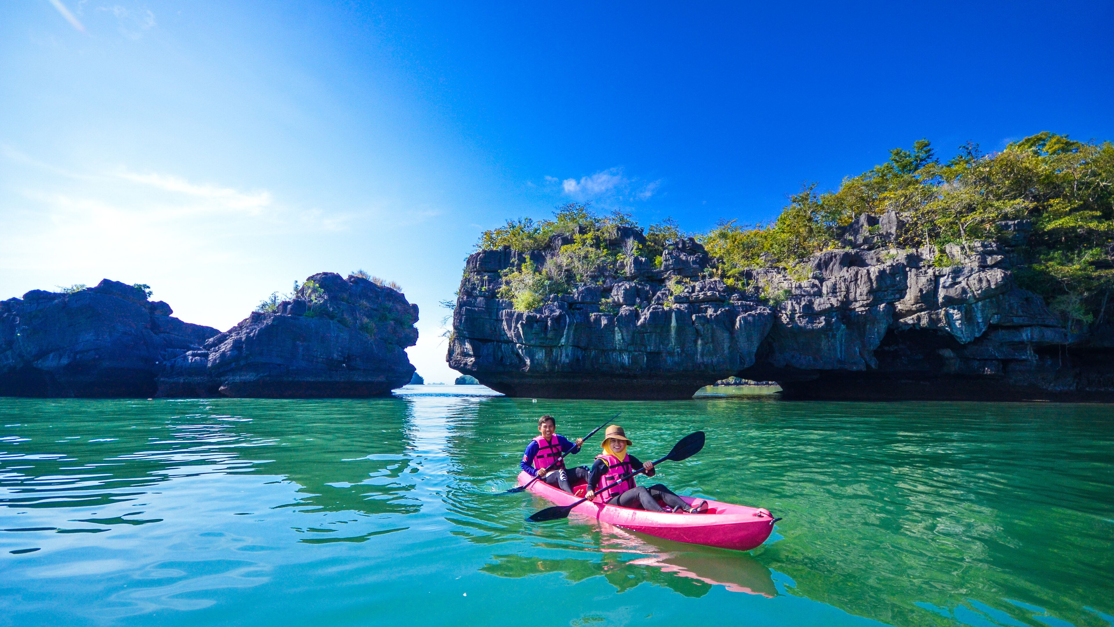 Two people kayaking on clear water with rocky cliffs and a small island in the background.