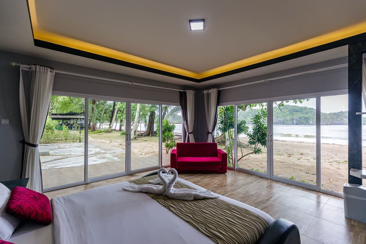 A hotel room with a large bed, a red chair, and a view of a beach and water.