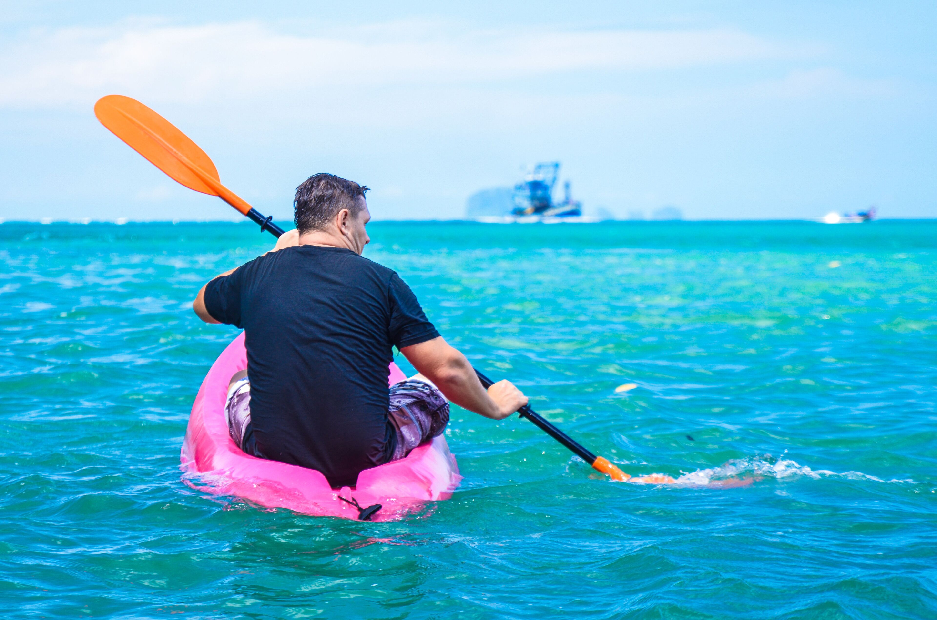 Una persona practica kayak en un bote inflable rosa en el océano, con un barco al fondo.