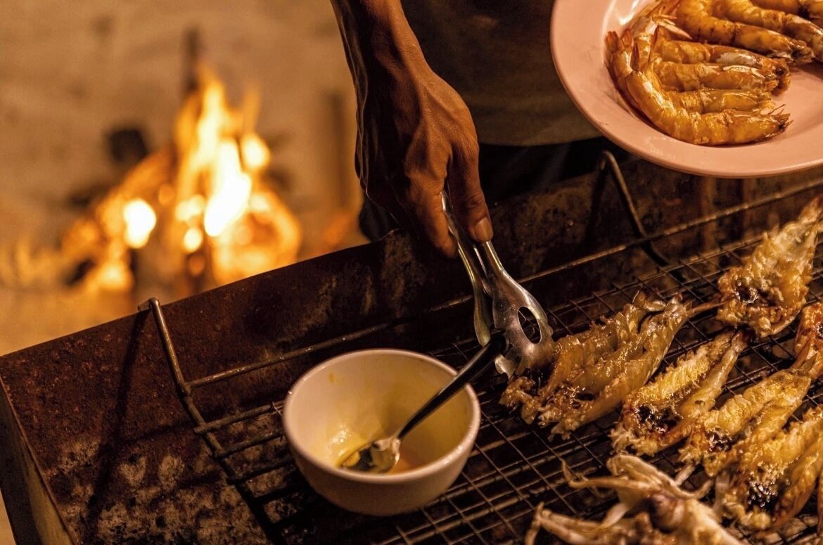 A person grilling seafood on a grill with a bowl of sauce nearby.