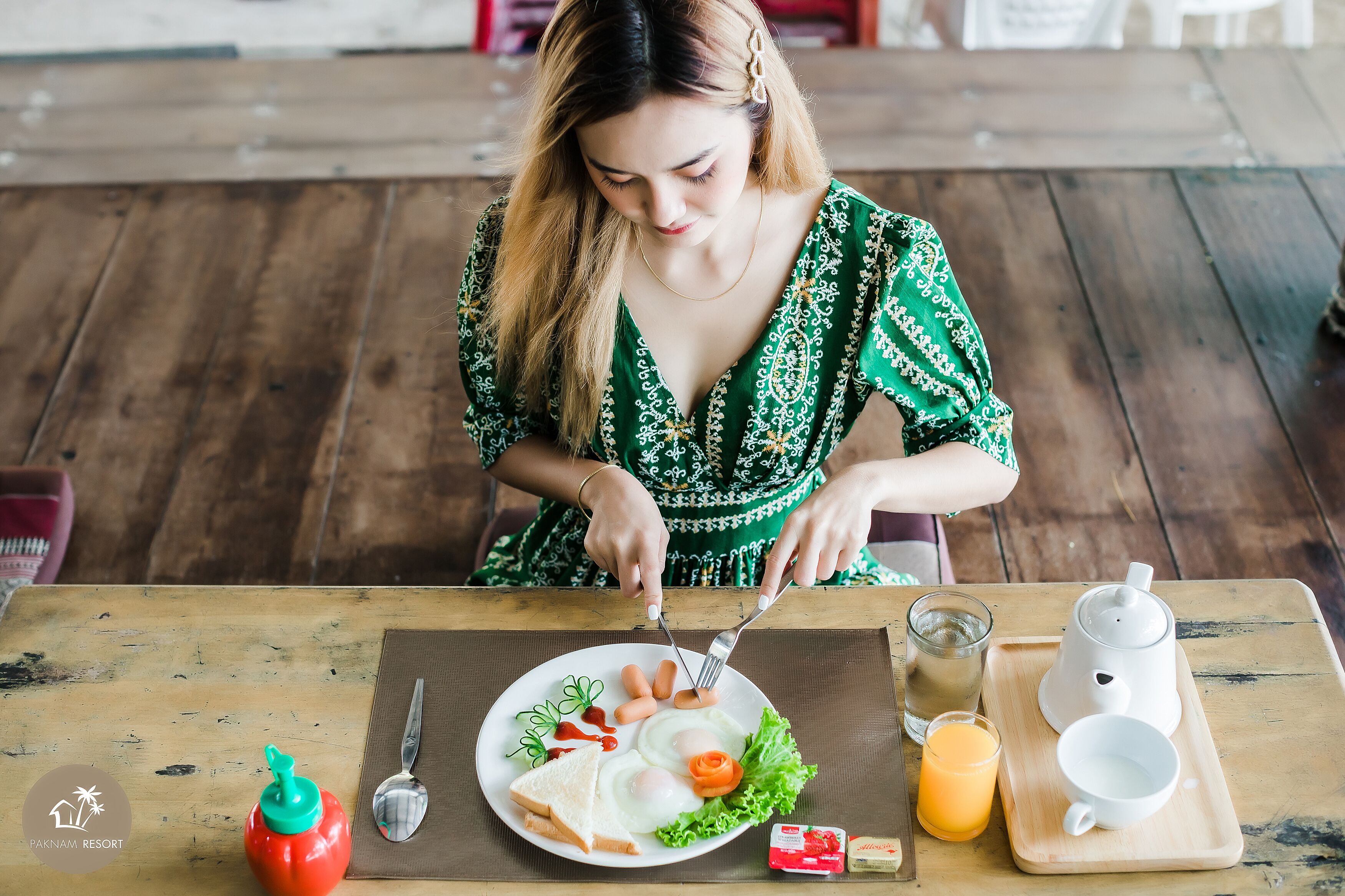 A person sitting at a wooden table with a plate of food, a glass of juice, and a teapot.