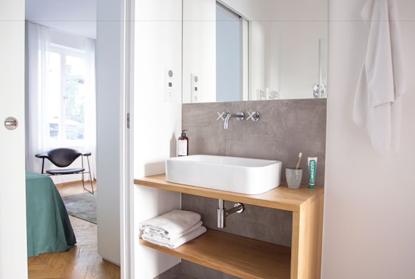 A modern bathroom with a large white sink, a mirror, and a wooden shelf with towels. - August The Boardinghouse (Hamburg)