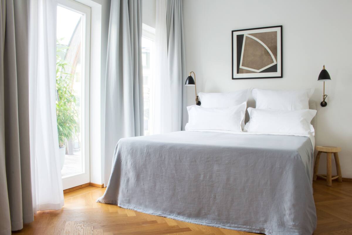 A neatly made bed with a grey bedspread, two bedside lamps, and a framed abstract artwork on the wall.