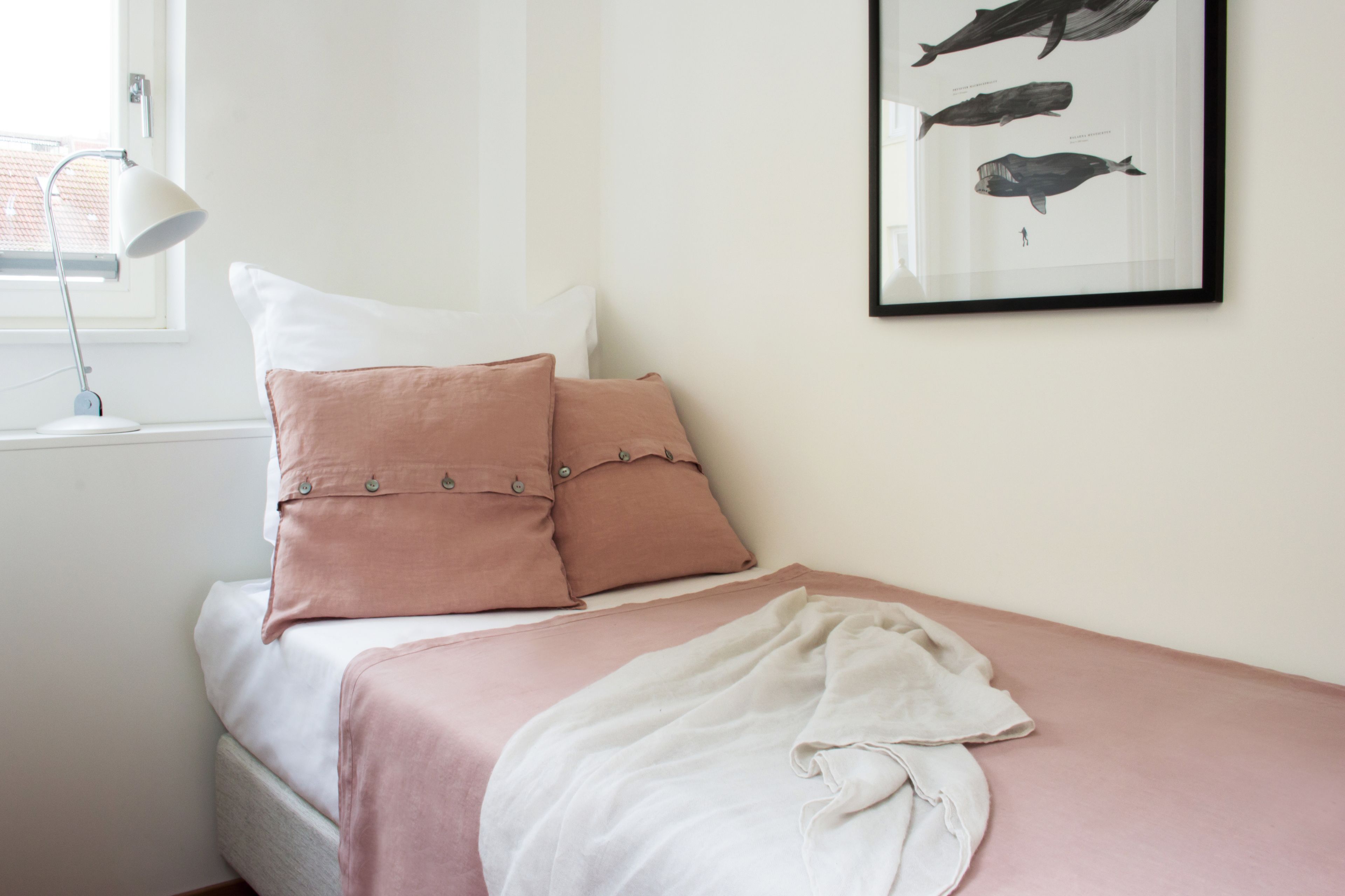 Un dormitorio con cama, almohadas, una manta, un cuadro colgado en la pared con ballenas y una ventana con lámpara.