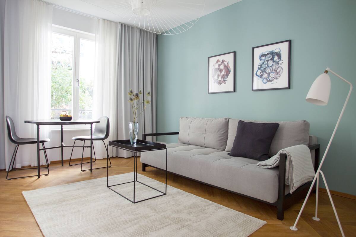A modern living room with a sofa, a coffee table, and a dining area with chairs. There are two framed artworks on the wall and a floor lamp.