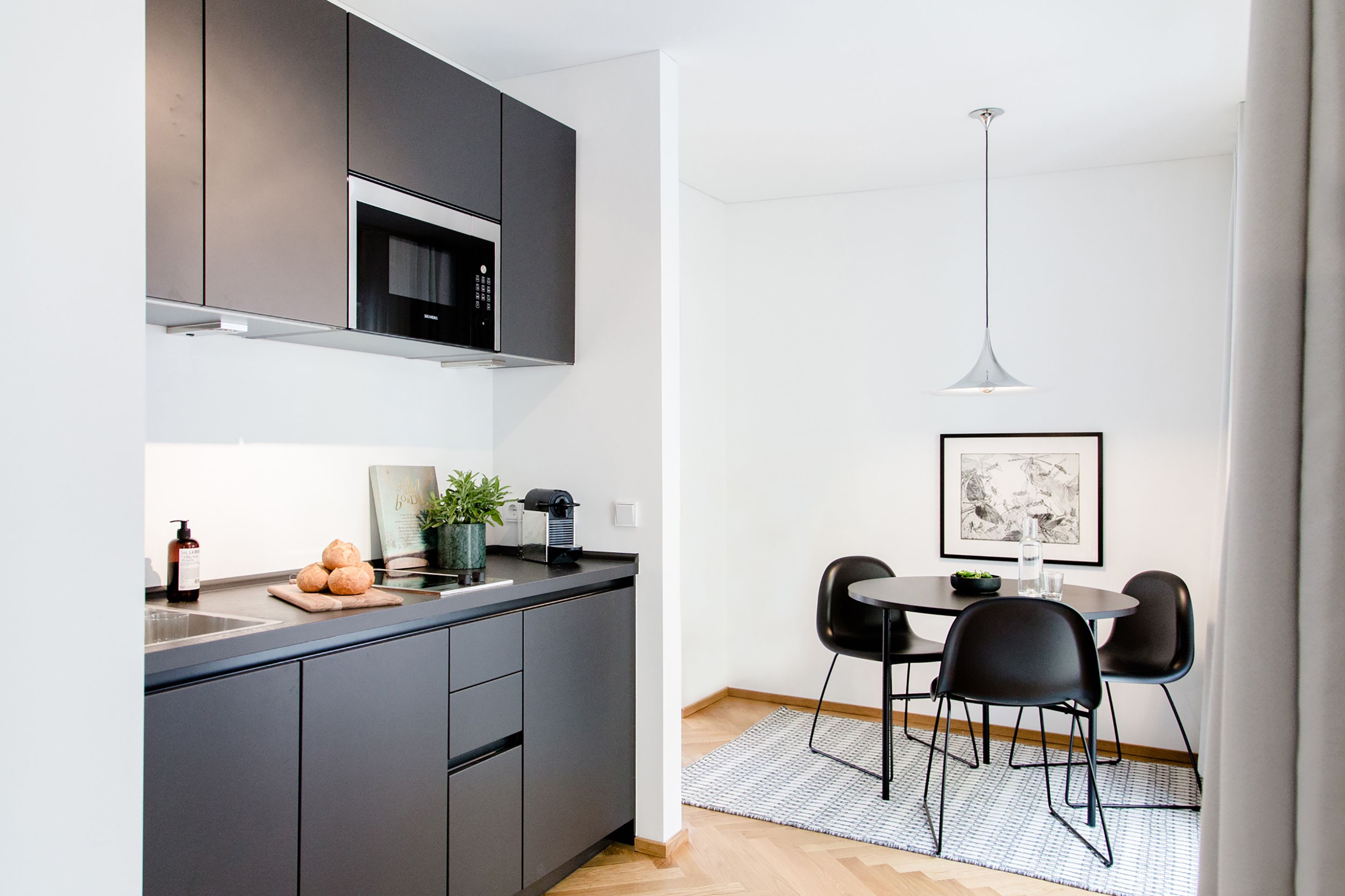 A modern kitchen with dark cabinets, a microwave, and a coffee machine. There is a dining area with a round table and black chairs.