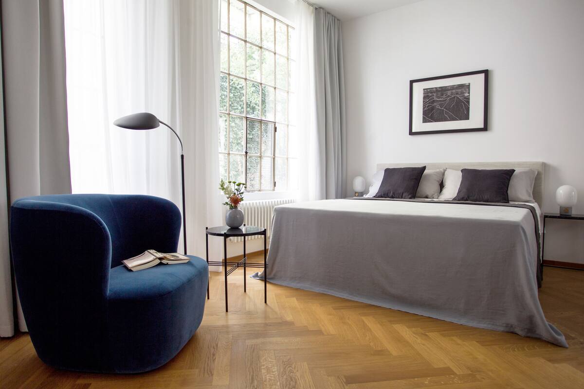 A modern bedroom with a large bed, a blue armchair, a small table with a vase, and a window with sheer curtains.