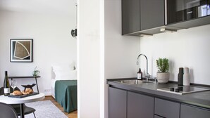 Cocina moderna con armarios grises, un lavamanos de acero inoxidable y un horno empotrado. Hay un pequeño comedor con una mesa y sillas, y una planta en maceta sobre la encimera.